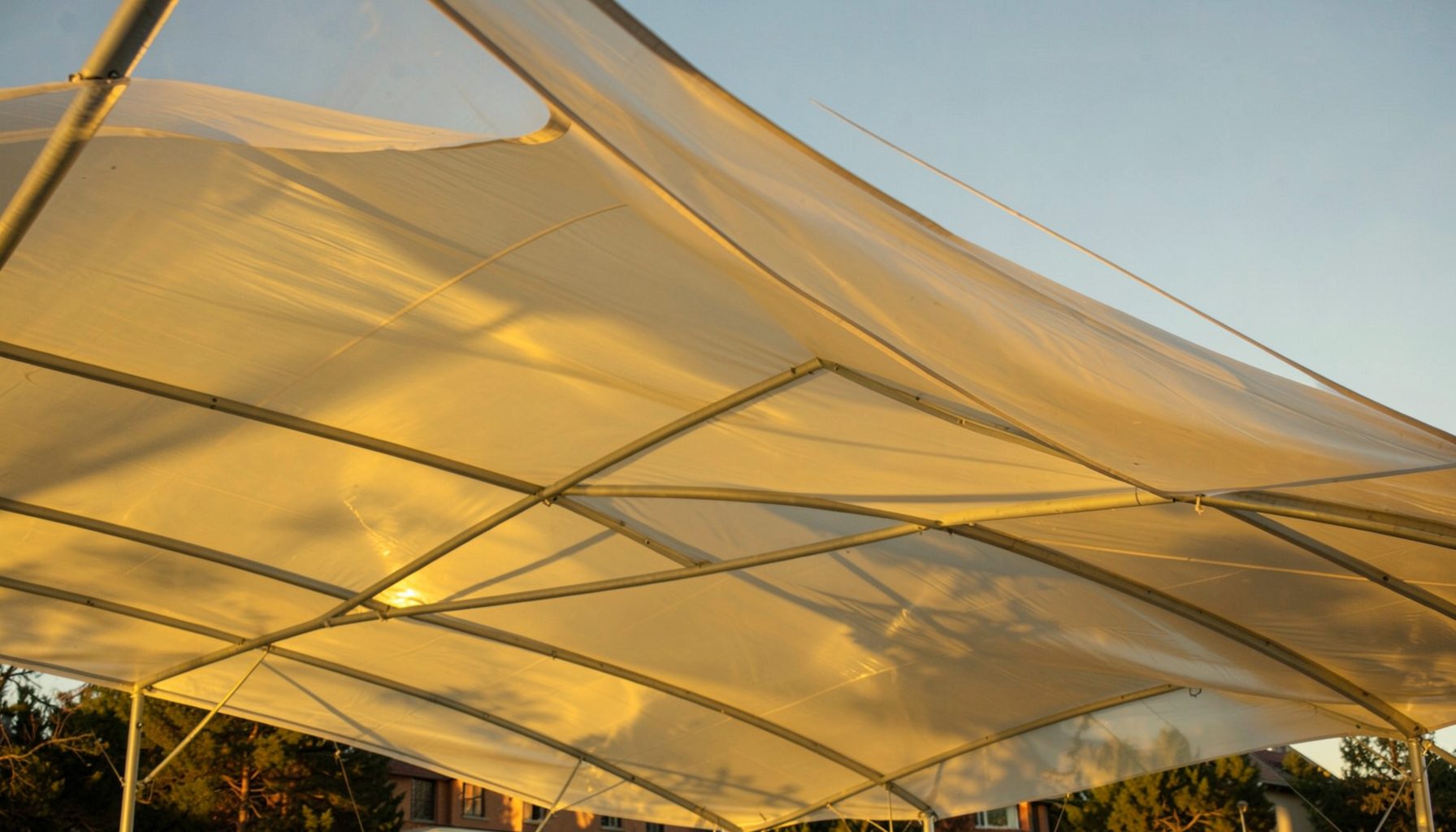 Sweeping tensile fabric roof structure made of translucent PTFE membrane material, illuminated by warm golden-hour sunlight against a clear sky.